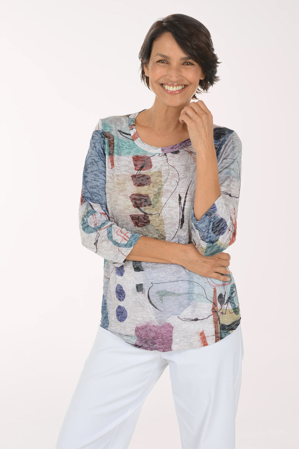 A smiling woman wearing a three-quarter sleeve top with an abstract multicolor print and white pants, standing against a plain white studio background.