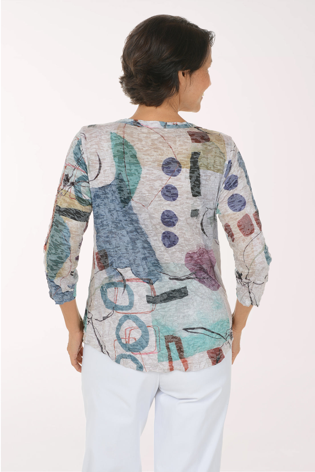 A smiling woman wearing a three-quarter sleeve top with an abstract multicolor print and white pants, standing against a plain white studio background.