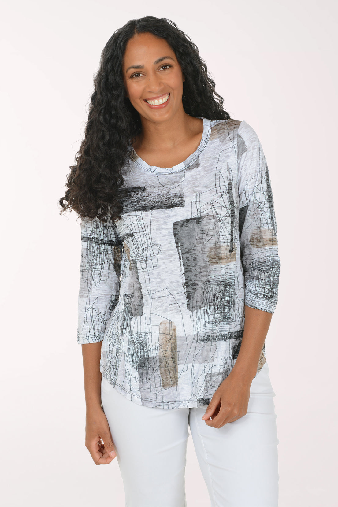 A smiling woman with long curly black hair wearing a three-quarter sleeve abstract print top in white, gray, and black, paired with white pants, against a plain light background.