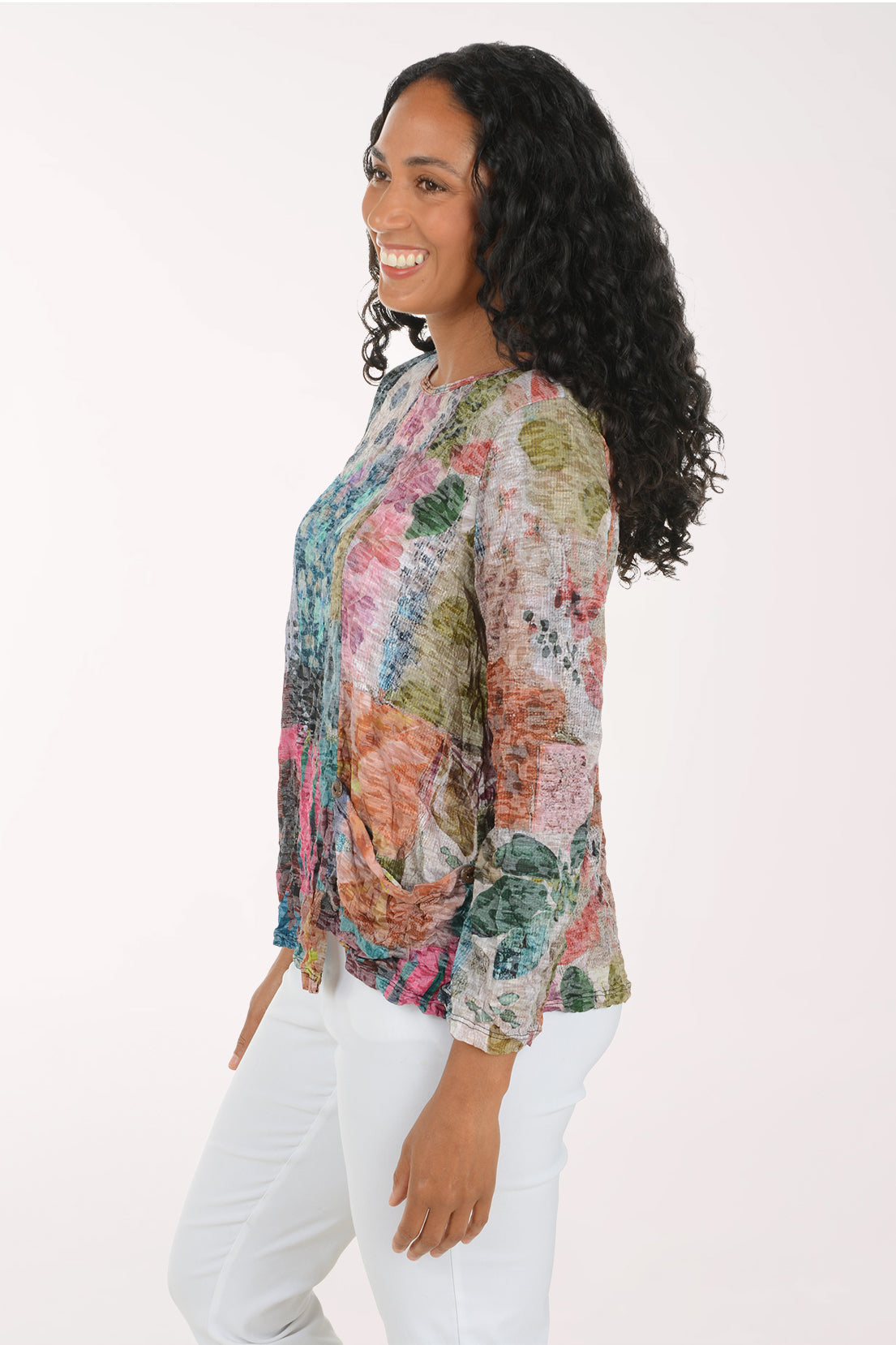 A woman with long, curly black hair is shown from a side-back angle, wearing a long-sleeve blouse with a multicolor abstract floral print and white pants, set against a plain white studio background.