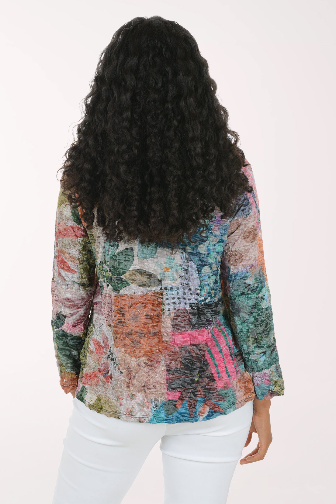 A woman viewed from behind wearing a long-sleeve patchwork print blouse with white pants, set against a plain off-white background.