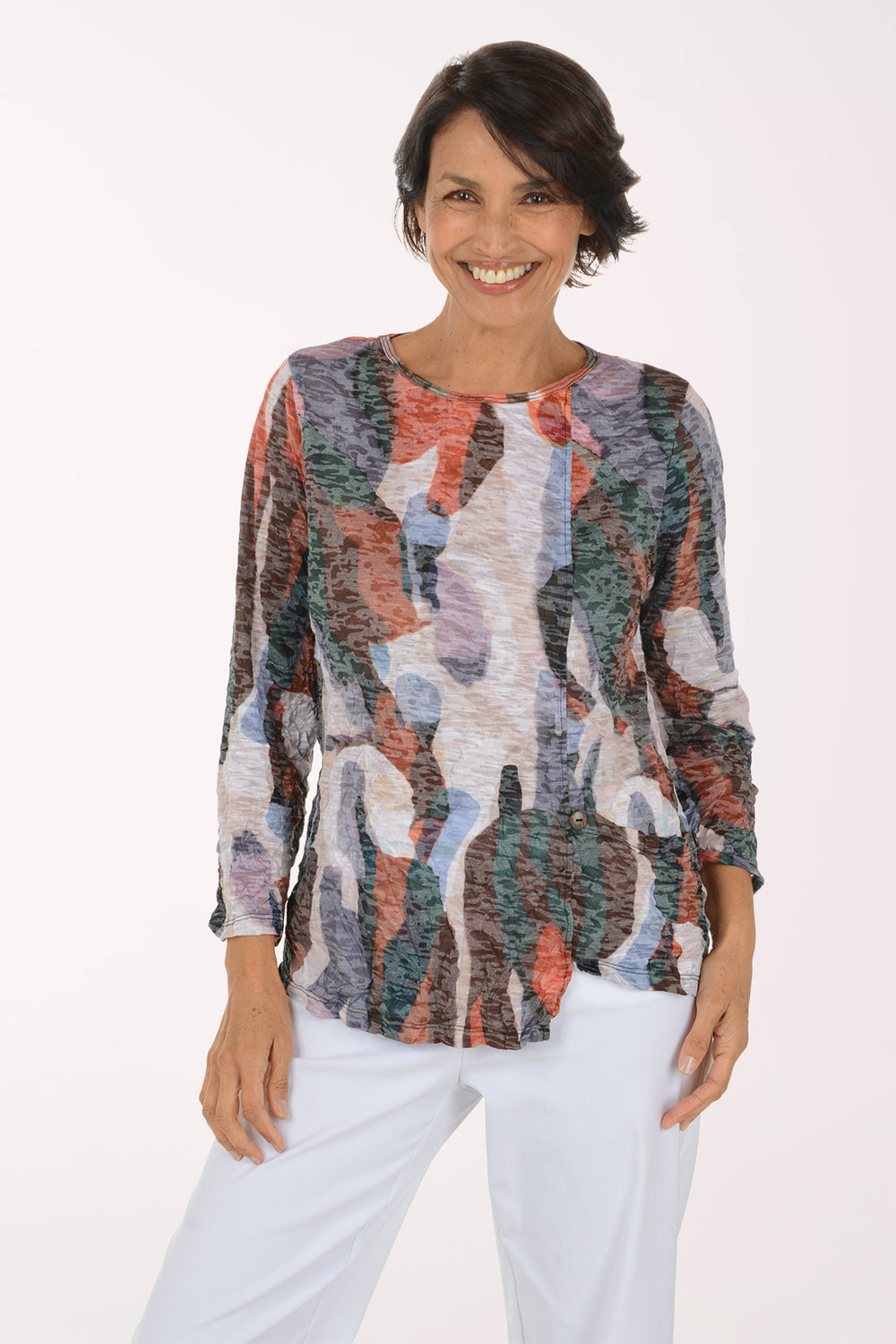 A smiling woman wearing a long-sleeve abstract print top with a mix of orange, blue, green, gray, and white tones, paired with white pants, against a plain white studio background.
