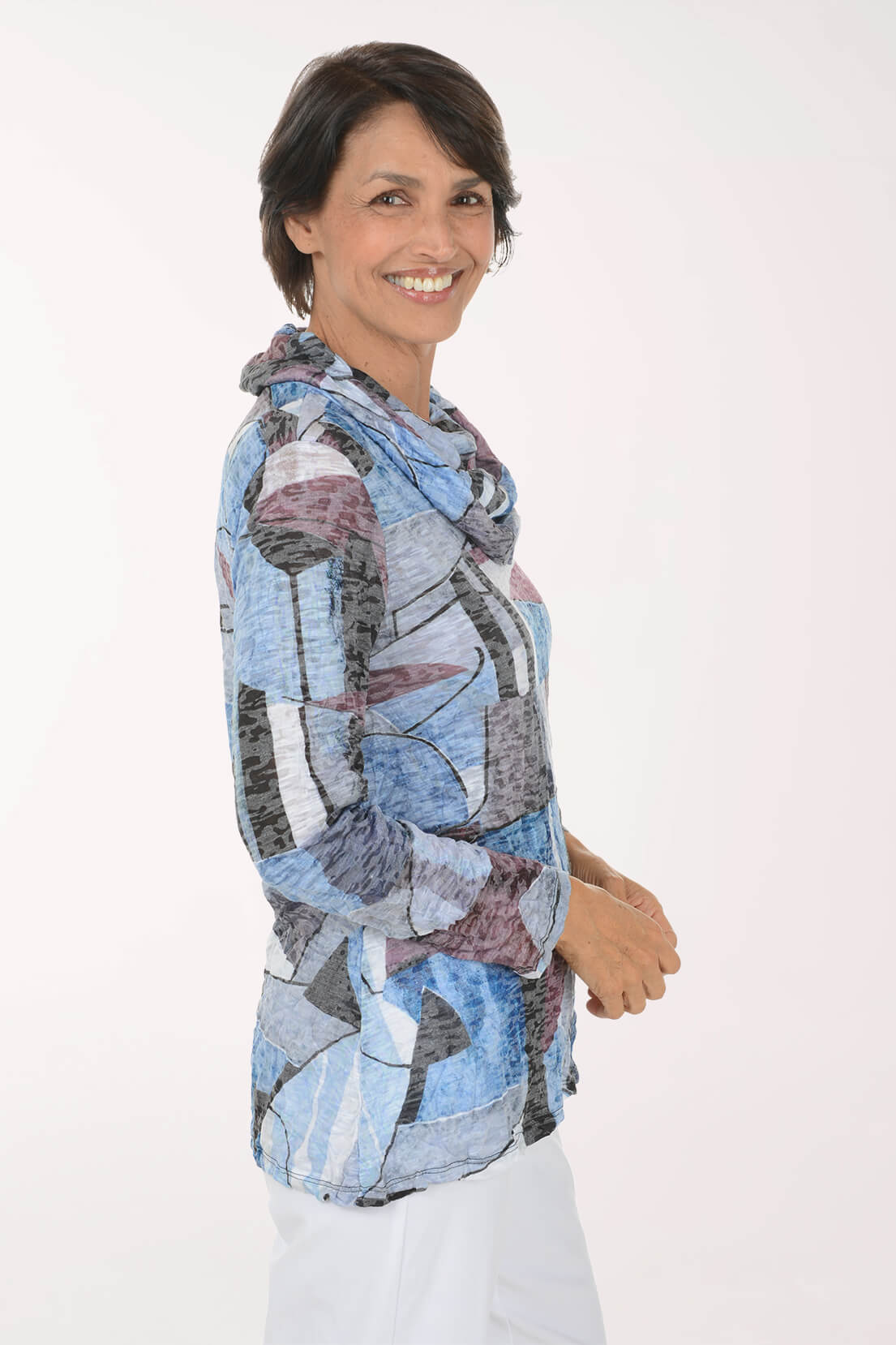 A smiling woman with short dark hair is shown from the waist up, wearing a lightweight hooded top with an abstract patchwork print in light blue, gray, white, and muted mauve. She is standing against a plain white studio background.