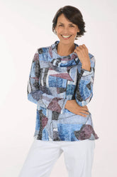 A smiling woman wearing a long-sleeve abstract patterned top with blue, gray, black, and mauve geometric shapes, paired with white pants, against a plain white studio background.