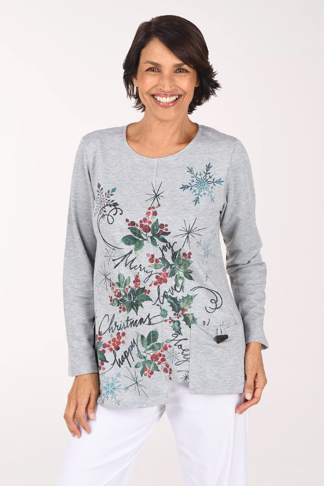 Smiling woman in a heather grey long sleeve scoop neck fleece top with a front graphic of red holly berries, green leaves, blue snowflakes and black holiday script, with a stepped asymmetrical hem and white pants