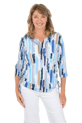 Woman wearing a blue and white patterned blouse with white pants on a white background.