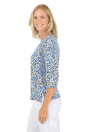 Woman wearing a blue and yellow patterned top with white pants on a white background. Side shot.