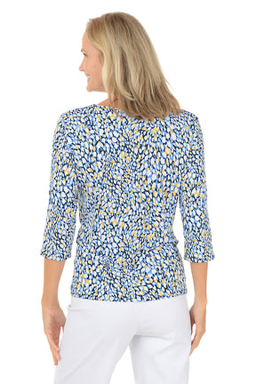 Woman wearing a blue and yellow patterned top with white pants on a white background. Back shot.