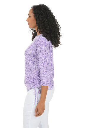 Woman wearing a purple paisley patterned top and white pants on a white background. Side shot.