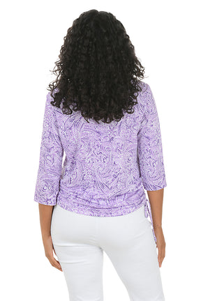 Woman wearing a purple paisley patterned top and white pants on a white background. Back shot.