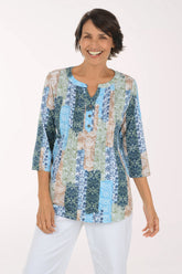 A smiling woman wearing a three-quarter sleeve patchwork top with a split neckline and white pants, photographed against a plain light background.