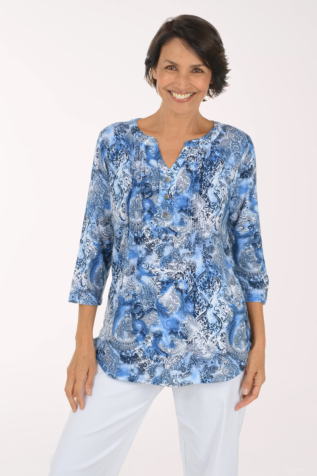 A woman modeling a blue and white abstract patterned long-sleeve top with a split neckline, paired with white pants, against a plain white studio background.