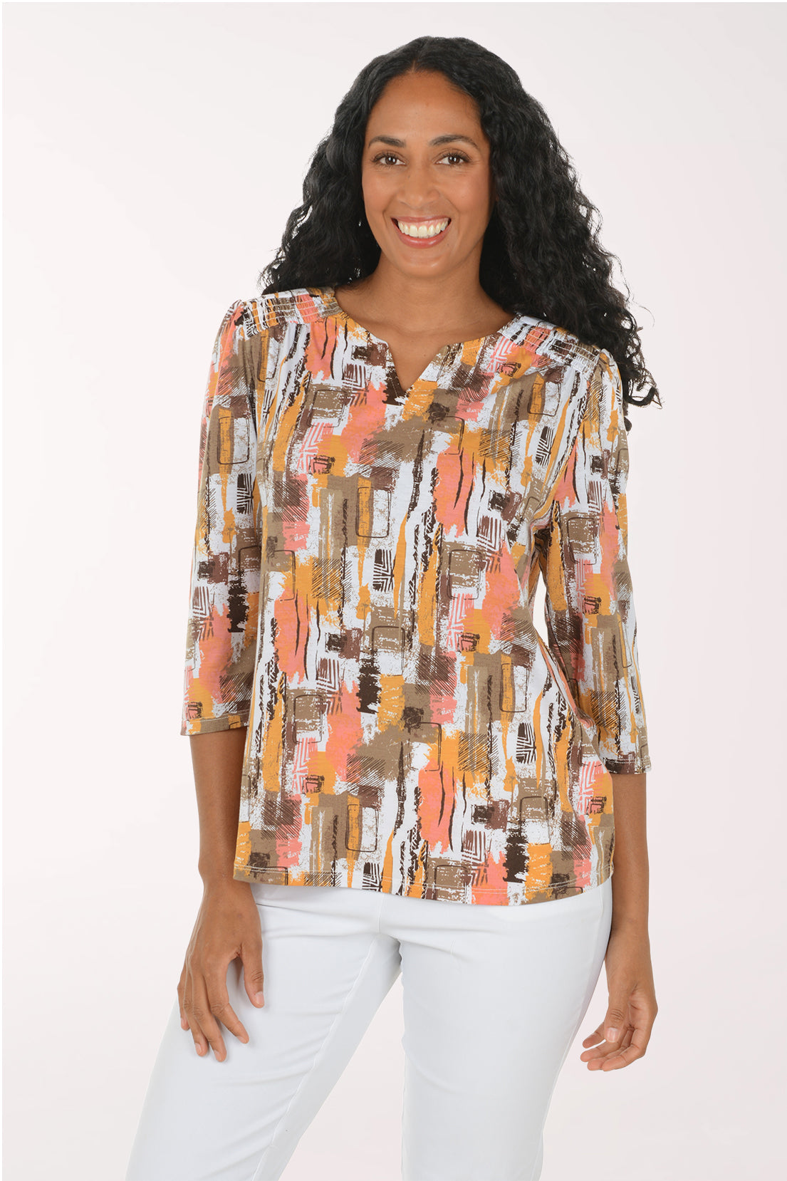 A smiling woman with long curly black hair models a three-quarter sleeve blouse with an abstract brushstroke pattern in orange, brown, beige, and white, paired with white pants against a plain white studio background.