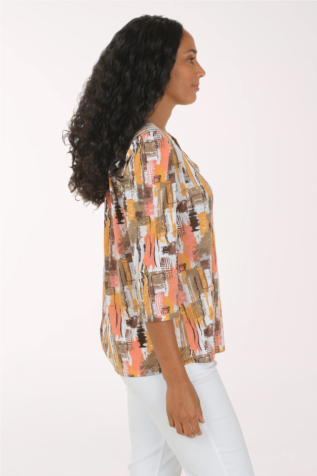 A smiling woman with long curly black hair models a three-quarter sleeve blouse with an abstract brushstroke pattern in orange, brown, beige, and white, paired with white pants against a plain white studio background.