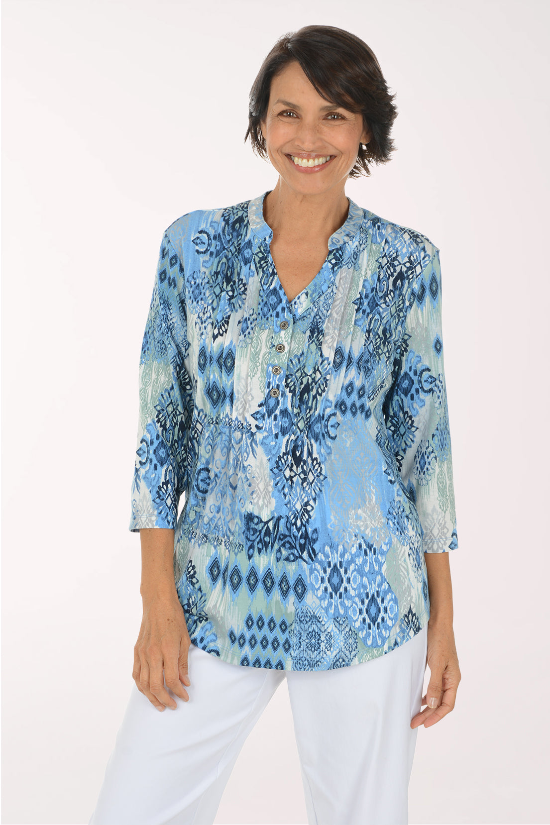A smiling woman wearing a light blue and white patterned blouse with three-quarter sleeves and white pants, standing against a plain white studio background.
