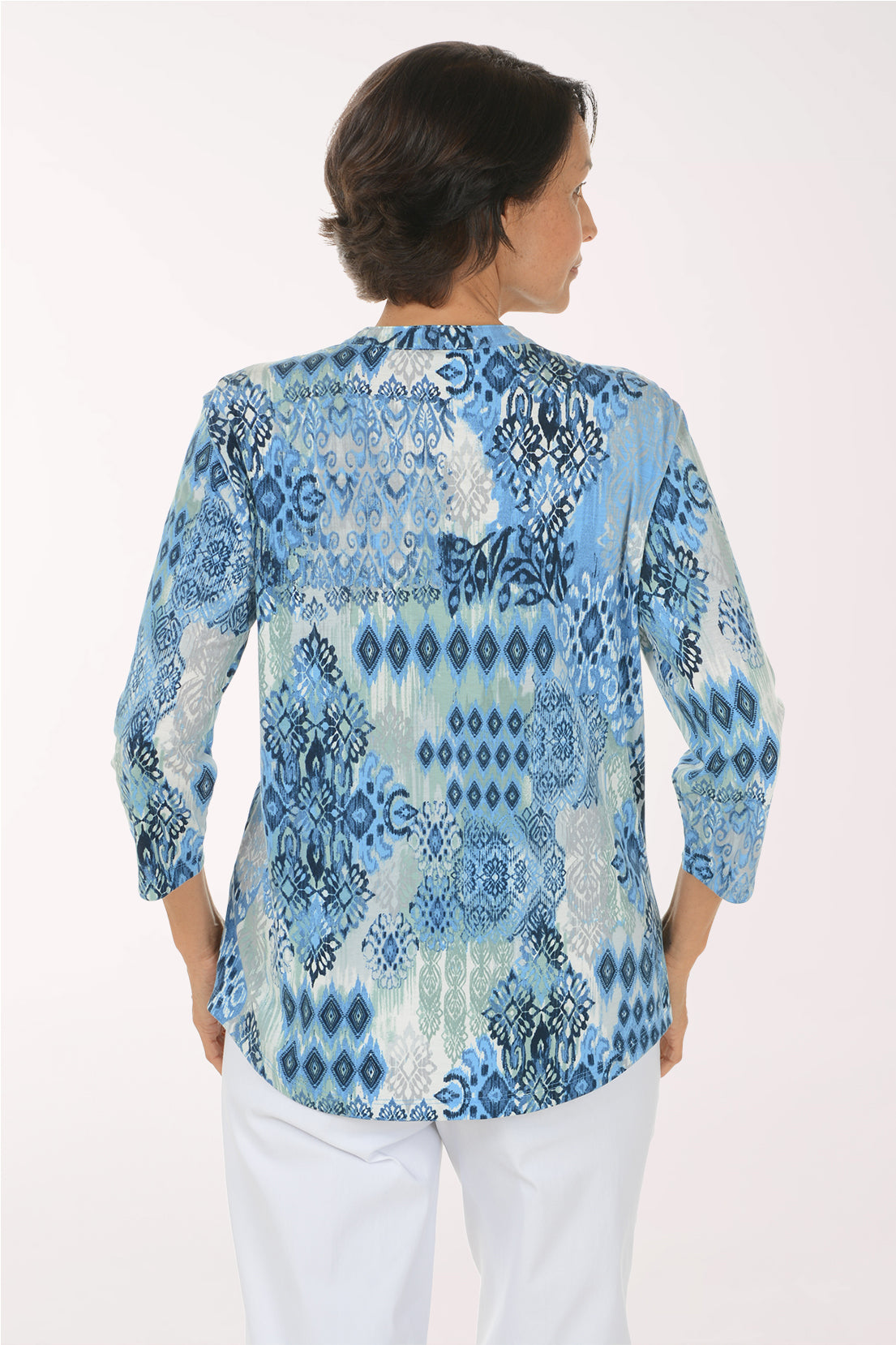 A smiling woman wearing a light blue and white patterned blouse with three-quarter sleeves and white pants, standing against a plain white studio background.