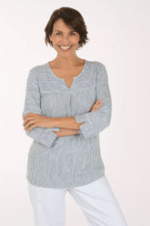 A smiling woman with short brown hair stands with arms crossed, wearing a light gray and white vertically textured long-sleeve top with a split neckline, paired with white pants, against a plain white studio background.