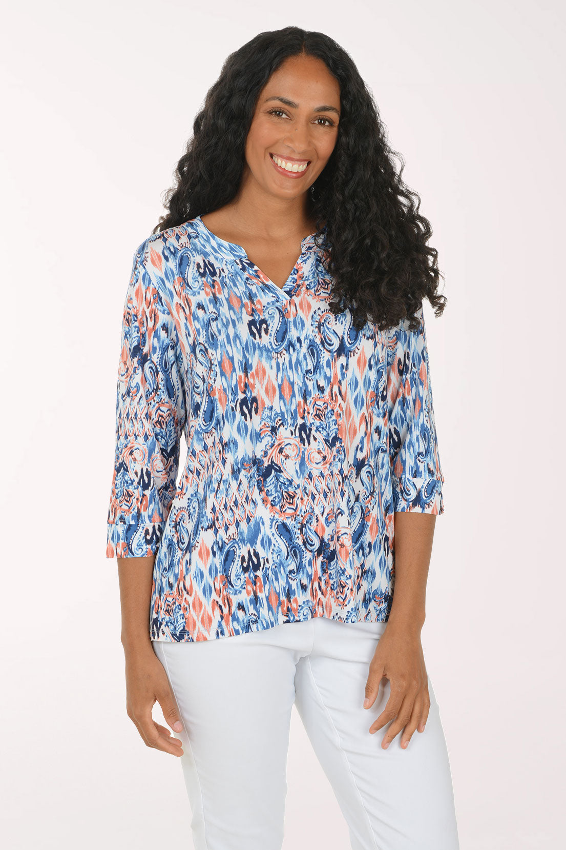 A smiling woman with long curly black hair models a three-quarter sleeve top with a blue, orange, and white abstract ikat pattern, paired with white pants, against a plain white studio background.