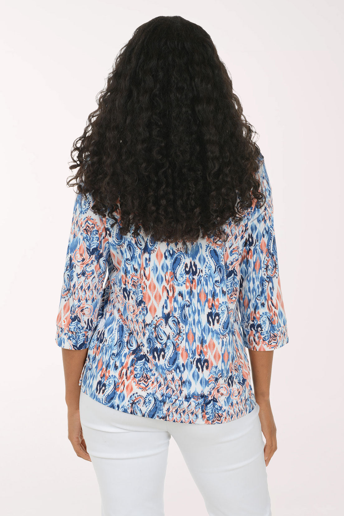 A woman with long curly black hair models a three-quarter sleeve top with a blue, orange, and white abstract ikat pattern, paired with white pants, against a plain white studio background.