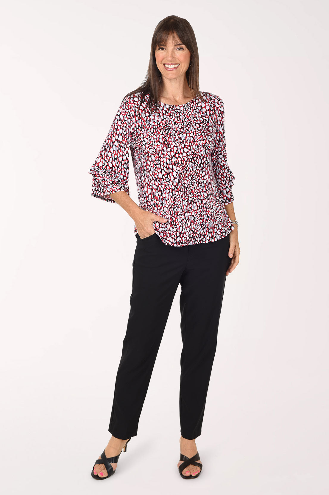 Full-length view of the black ankle pants with one hand in the pocket, styled with the spotted ruffle top and black heeled sandals.