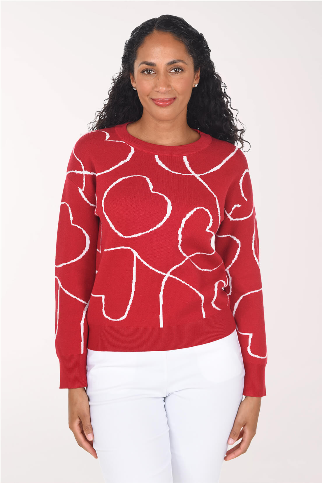 A woman wearing a red crewneck sweater with a white knit heart pattern, paired with white pants, standing against a plain light background.
