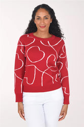 A woman wearing a red crewneck sweater with a white knit heart pattern, paired with white pants, standing against a plain light background.