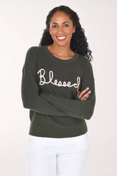 Front view of a woman wearing a dark olive green crewneck sweater with "Blessed" in cream rope script across the chest, styled with white pants.