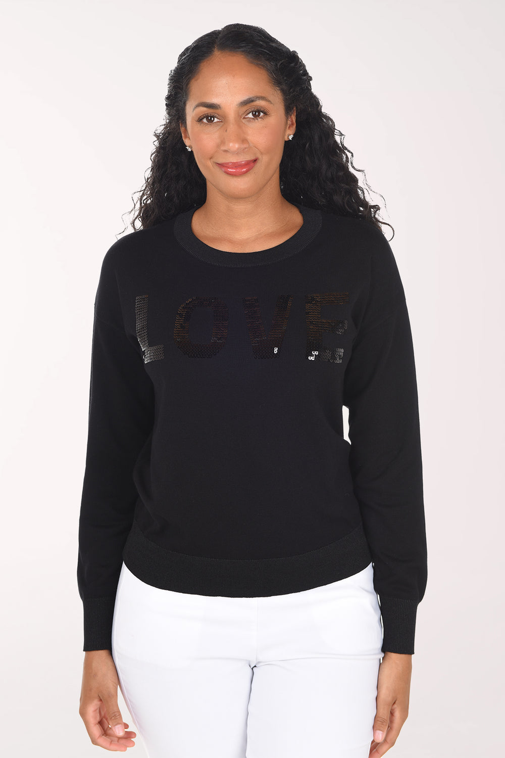Front view of the black long-sleeve sweater with black sequined "Love" lettering and ribbed cuffs and hem, worn with white pants.