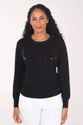 Front view of the black long-sleeve sweater with black sequined "Love" lettering and ribbed cuffs and hem, worn with white pants.