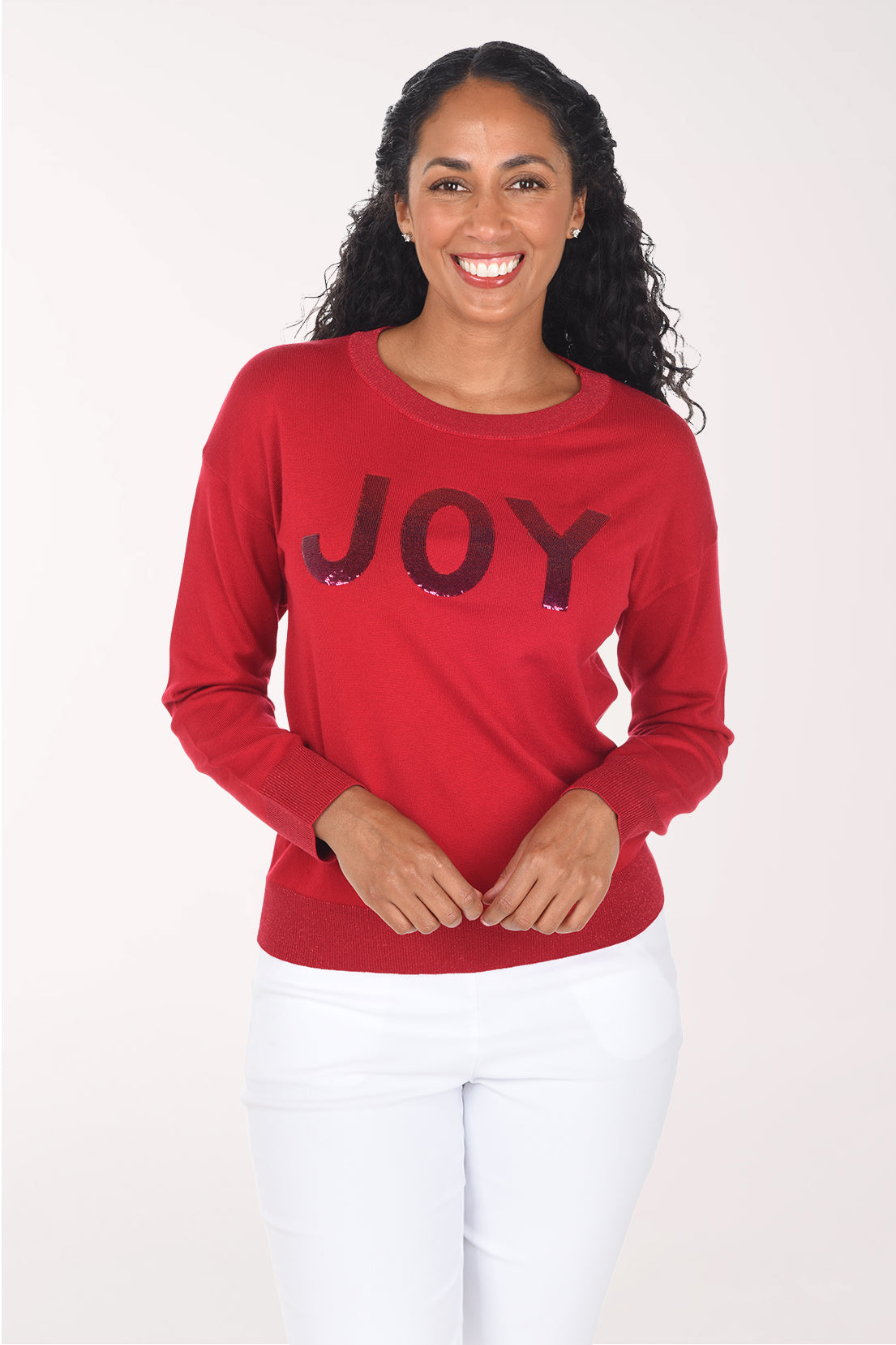 Front view of a woman wearing a red crewneck sweater with "JOY" in deep red sequined block letters, styled with white pants.