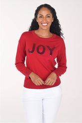 Front view of a woman wearing a red crewneck sweater with "JOY" in deep red sequined block letters, styled with white pants.