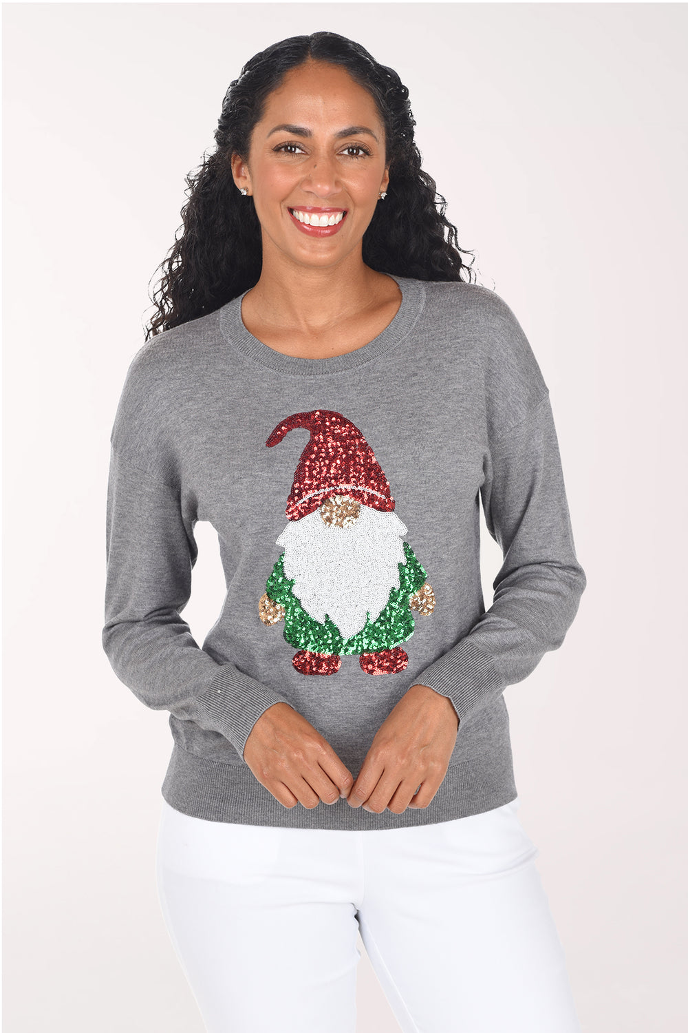 Front view of a woman wearing a heather grey crewneck sweater with a sequined holiday gnome on the front: red sequin hat, fuzzy white beard, gold nose and green sequin body, styled with white pants.