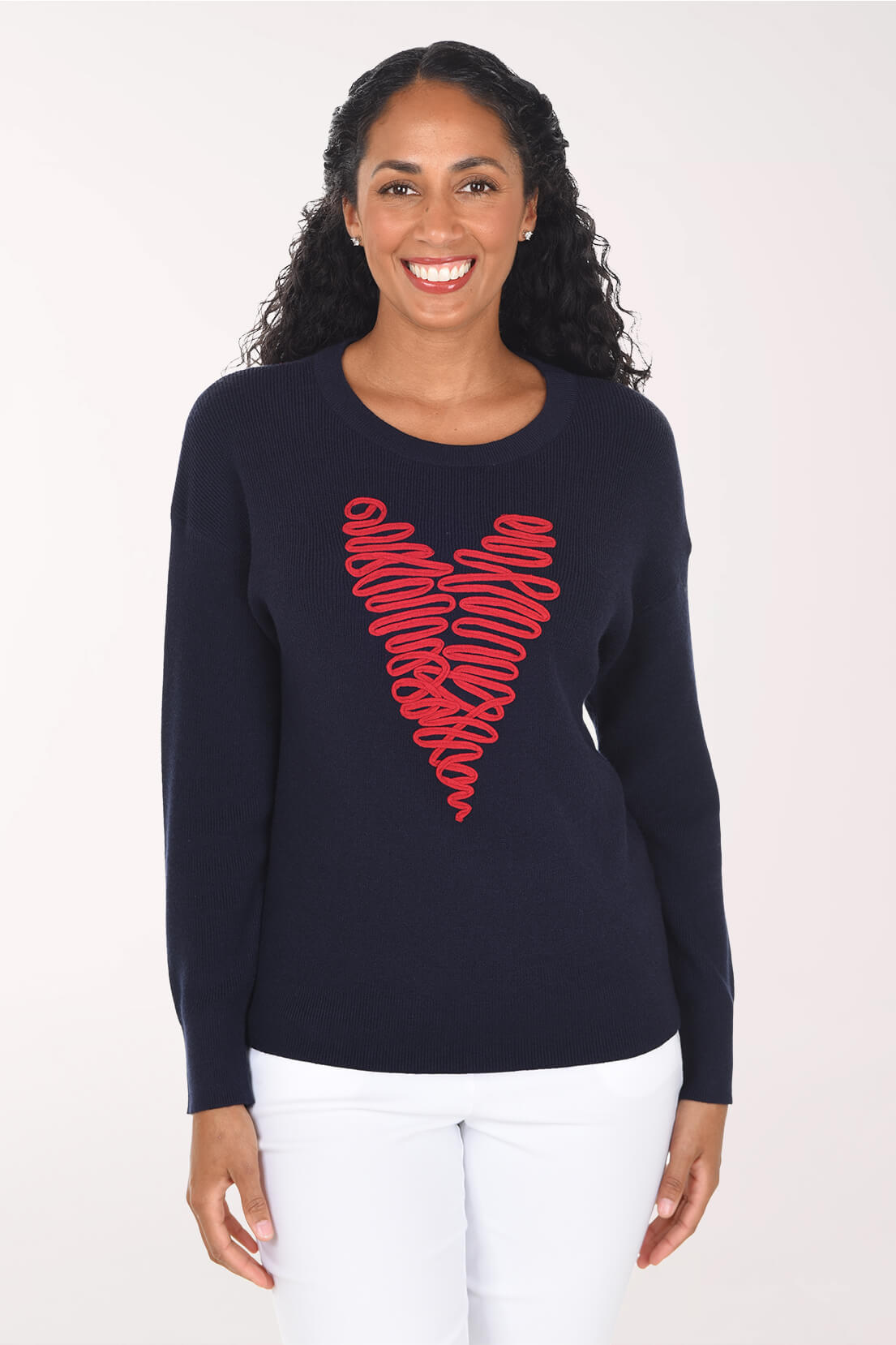 A smiling woman with long dark curly hair wears a navy blue long-sleeve knit sweater featuring a large red scribbled heart graphic on the front, paired with white pants, against a plain light background.