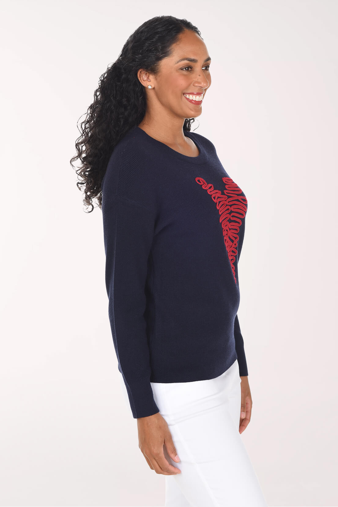 A woman wearing a navy blue crewneck sweater with a red zebra-striped heart graphic on the front, paired with white pants, photographed against a plain white studio background.