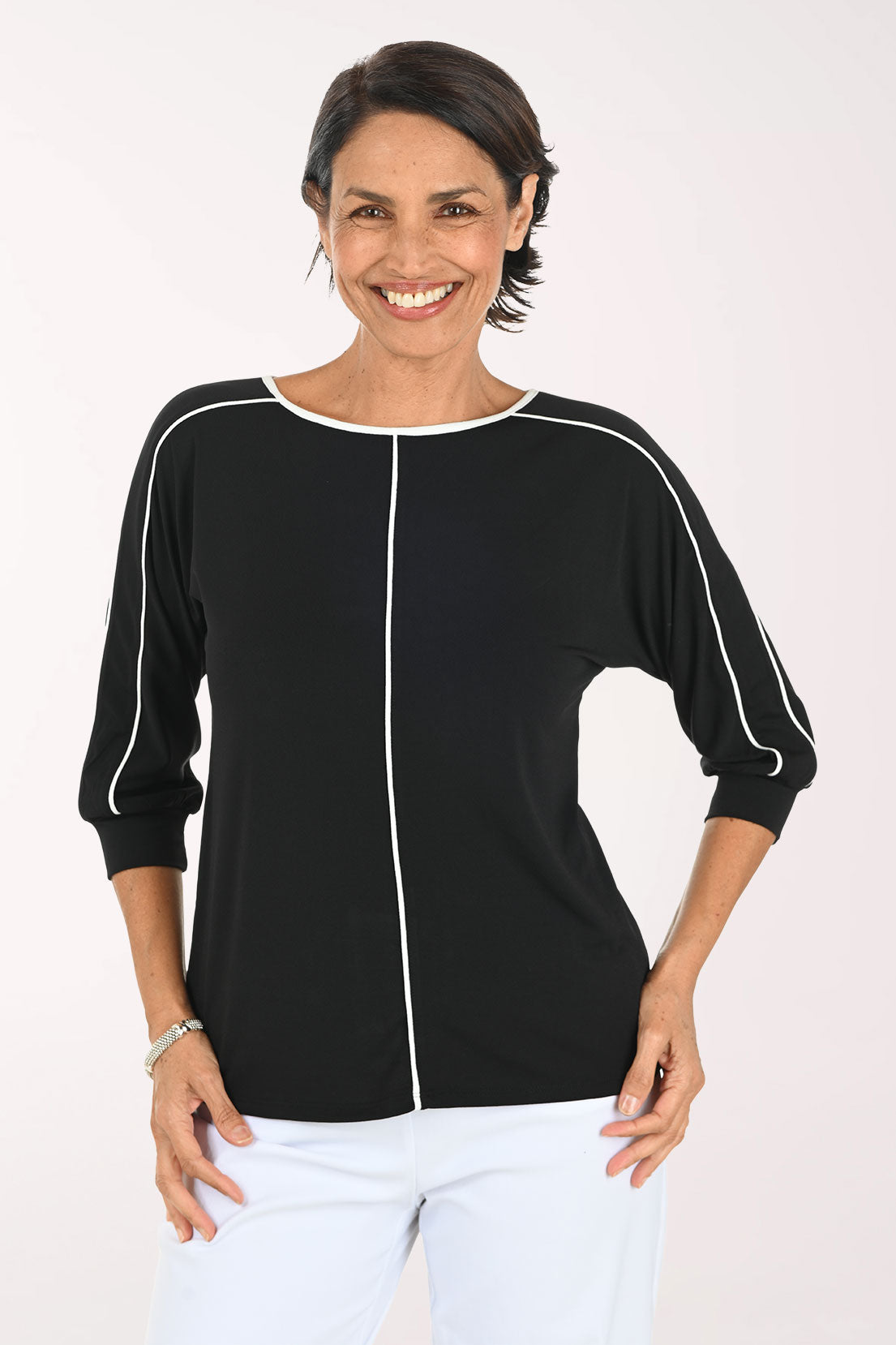 A smiling woman wearing a black three-quarter sleeve top with white contrast piping details, paired with white pants, against a plain light background.