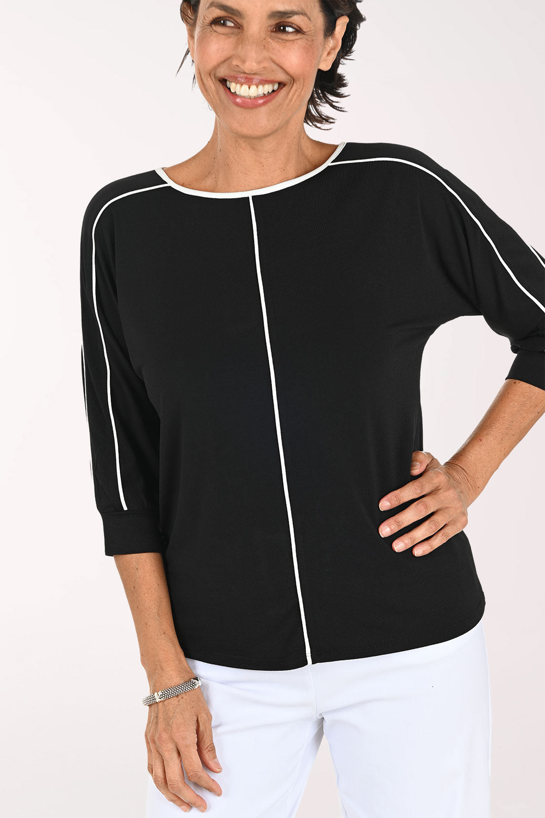 A smiling woman wearing a black three-quarter sleeve top with white contrast piping details, paired with white pants, against a plain white studio background.