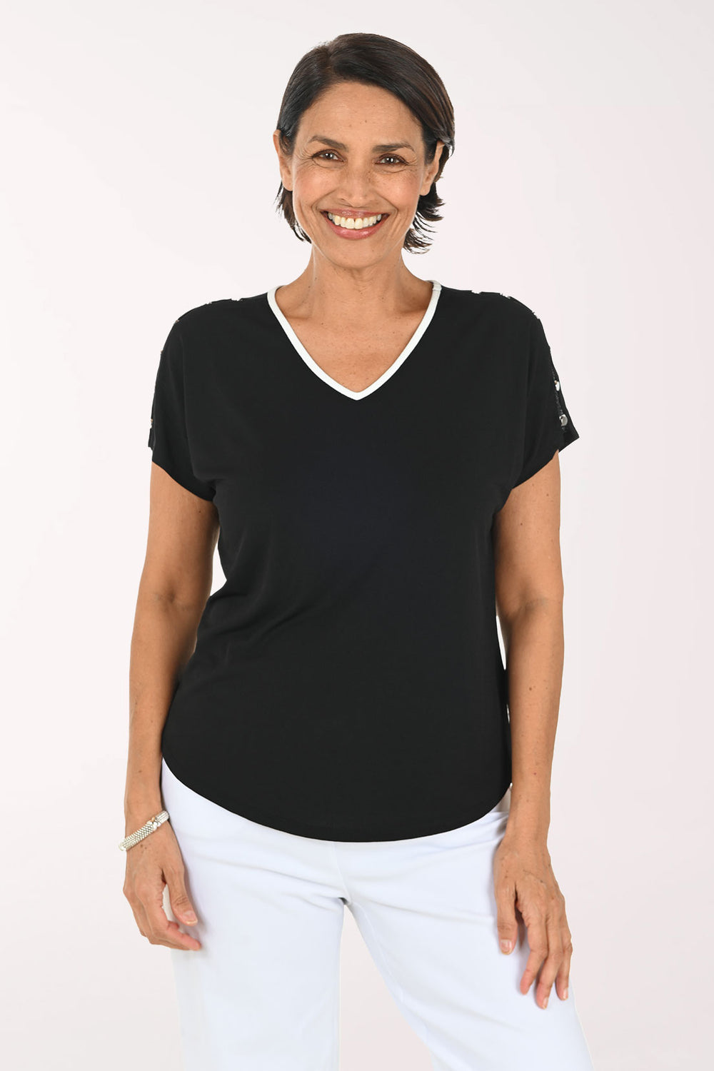 A smiling woman wearing a black short-sleeve V-neck top with a white trim and white pants, standing against a plain light background.