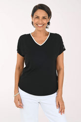 A smiling woman wearing a black short-sleeve V-neck top with a white trim and white pants, standing against a plain light background.