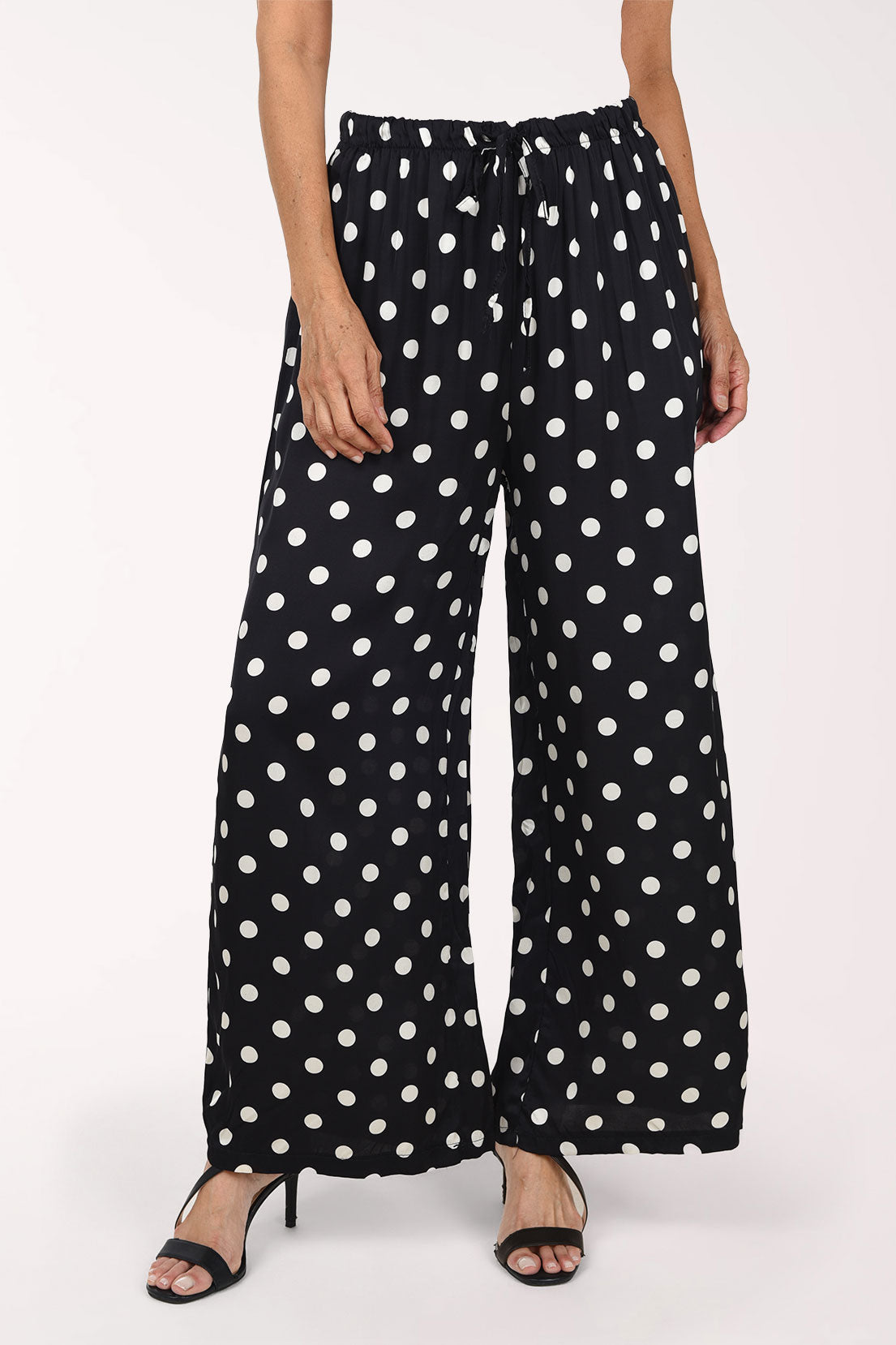 Black wide-leg pants with a white polka dot pattern, shown on a model wearing black strappy heels against a plain white studio background.