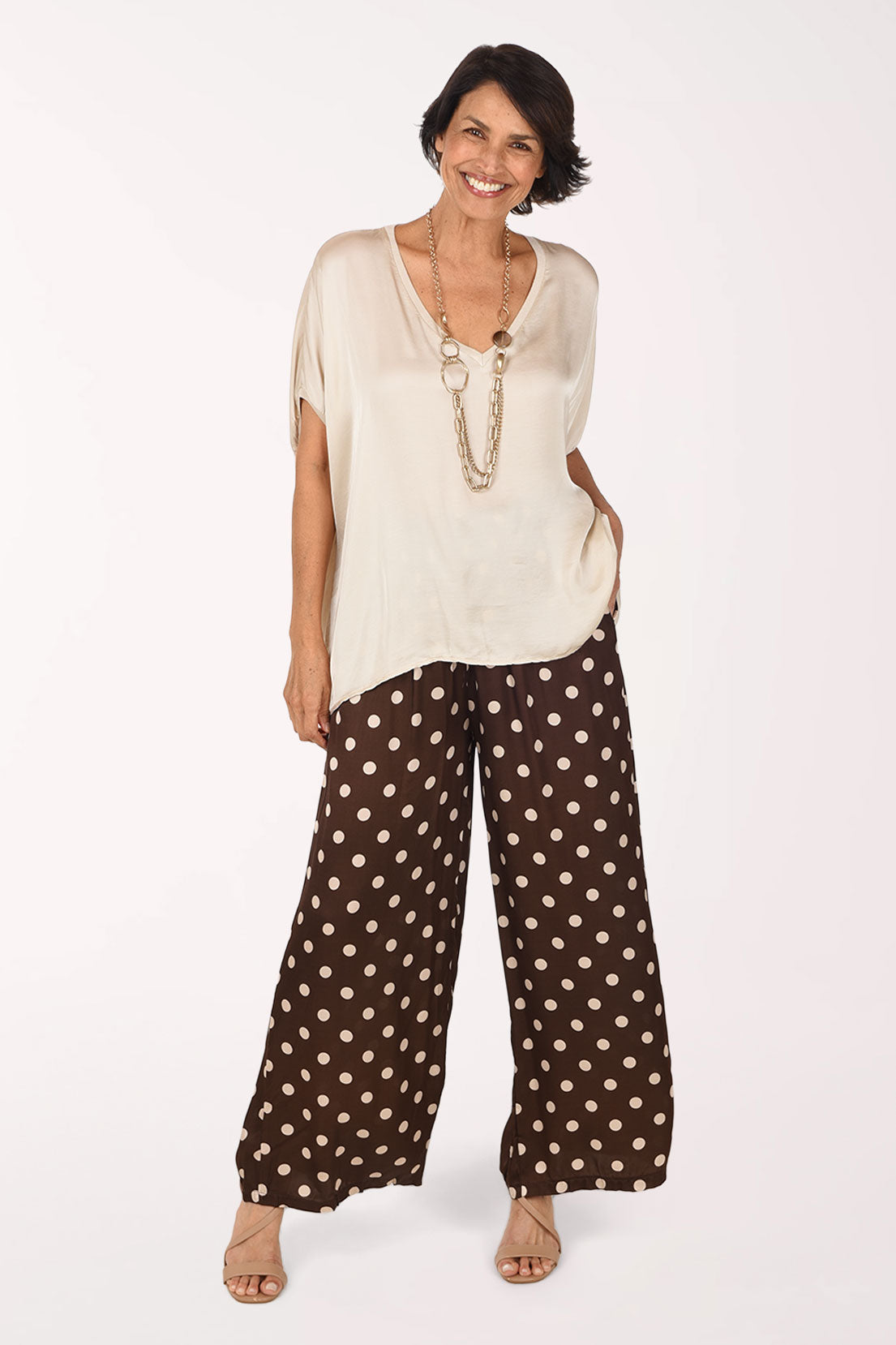 A woman modeling a cream-colored short-sleeve top and brown wide-leg pants with beige polka dots, standing against a plain white studio background.