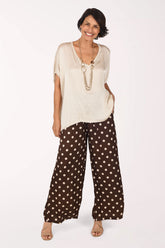 A woman modeling a cream-colored short-sleeve top and brown wide-leg pants with beige polka dots, standing against a plain white studio background.