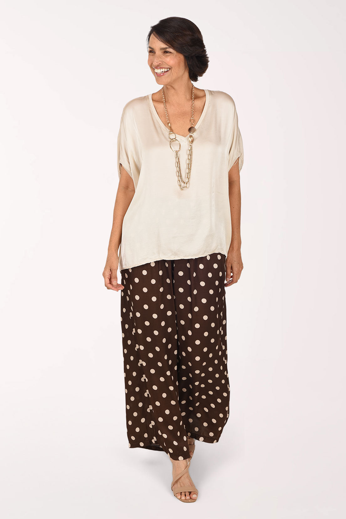 A woman modeling a cream short-sleeve top and brown wide-leg pants with beige polka dots, standing against a plain white studio background.