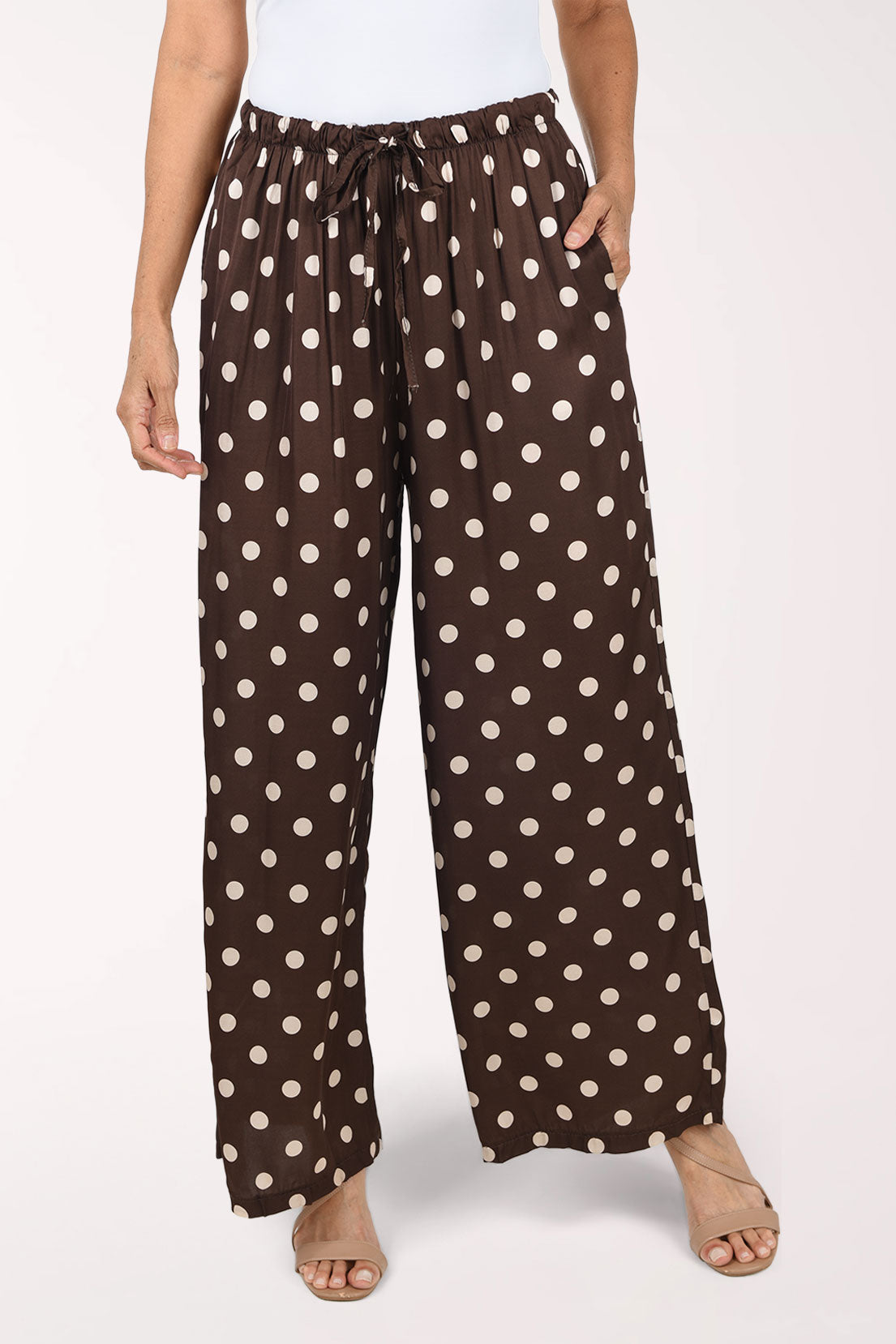 A person wearing loose brown wide-leg pants with a cream polka dot pattern, paired with a white top and beige sandals, shown against a plain light background.