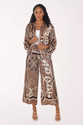A woman modeling a matching two-piece set in a brown and beige silk-like fabric with a white graphic print. The set includes a cropped jacket with a drawstring and wide-leg pants, styled with a white top and beige strappy heels against a plain white background.