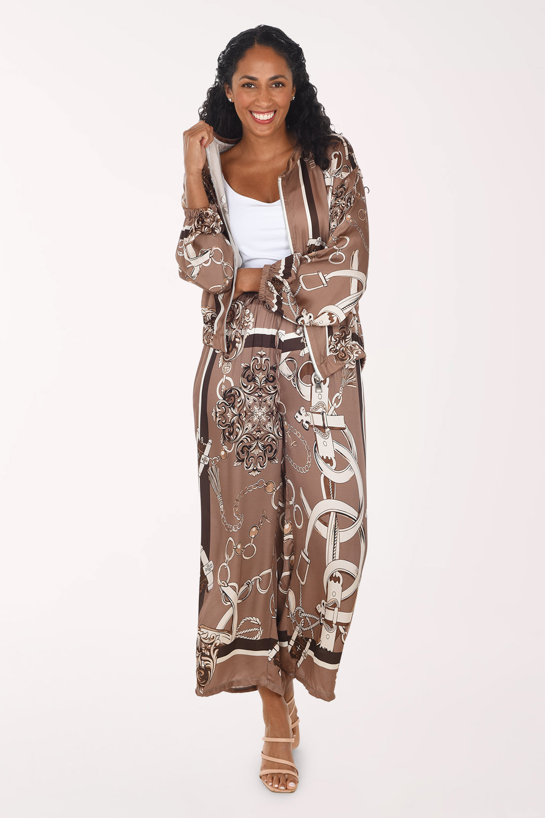 A woman modeling a matching brown and beige patterned set consisting of a long-sleeve zip-up hoodie and wide-leg pants, styled over a white crop top and tan heeled sandals against a plain white background.