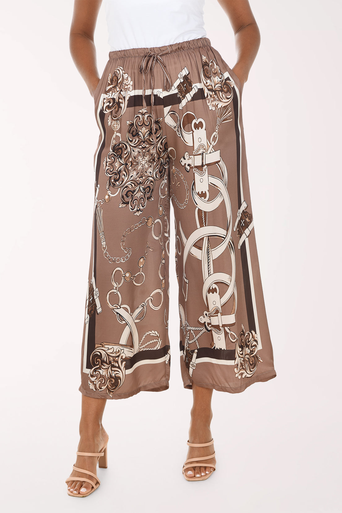 A model wearing brown wide-leg cropped pants with a white and beige chain-link and damask print, paired with a white top and beige heeled sandals, against a light studio background.