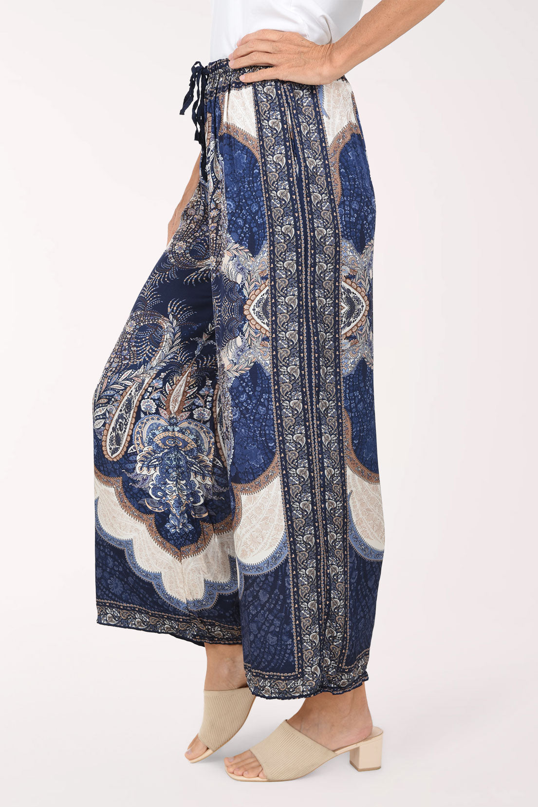 A person wearing loose, wide-leg cropped pants with a blue, beige, and white paisley and floral print, paired with beige heeled slide sandals against a plain light background.