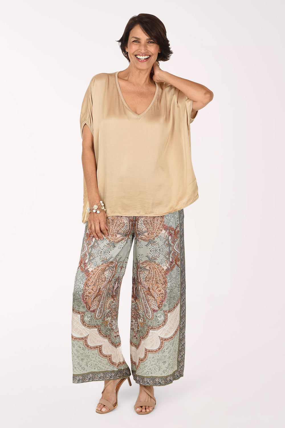 A woman modeling a beige short-sleeve V-neck top and wide-leg patterned pants with a paisley design in muted green, brown, and cream, set against a plain white studio background.