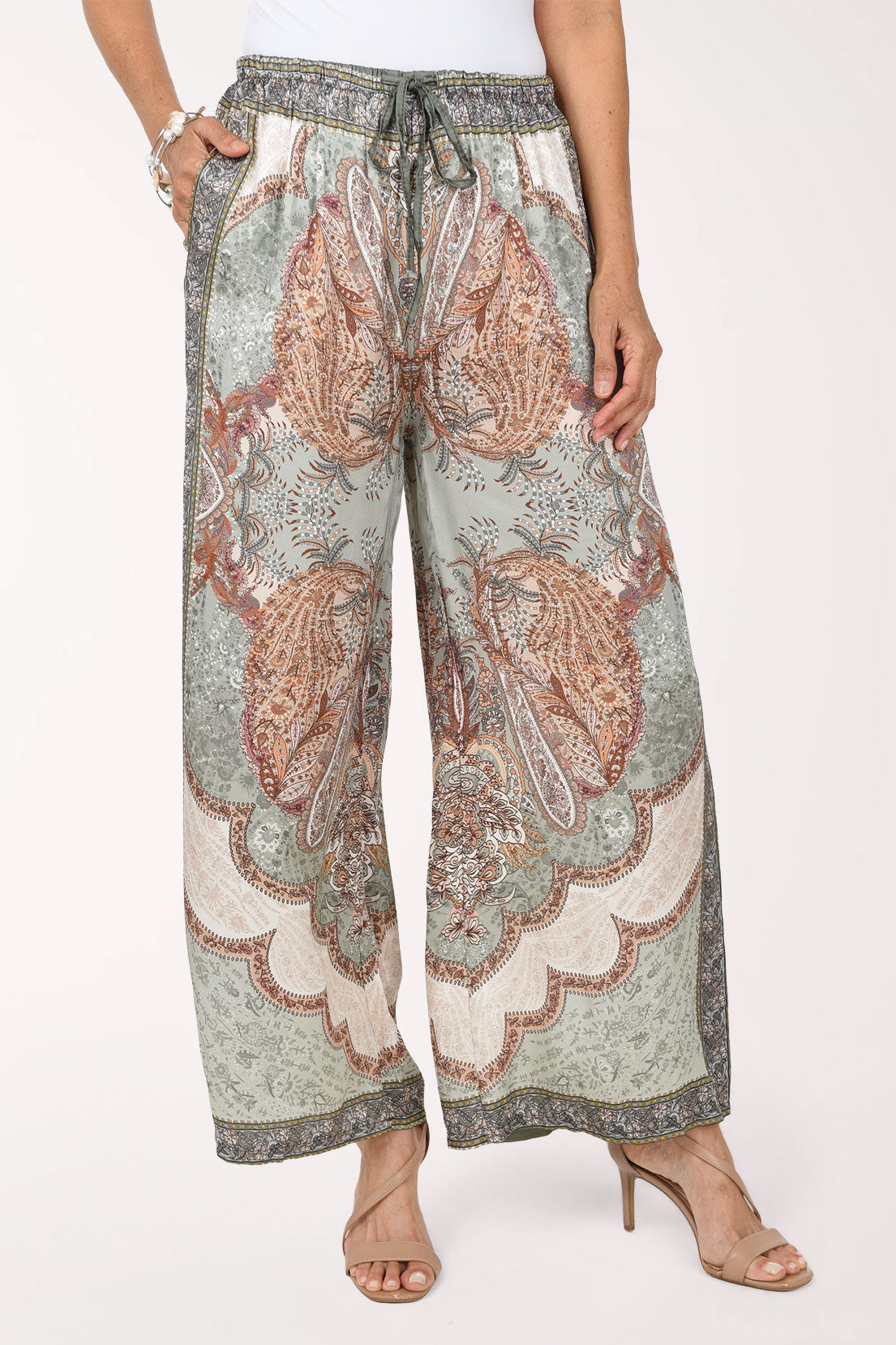 A woman modeling wide-leg patterned pants with a paisley design in muted green, brown, and cream, set against a plain white studio background.