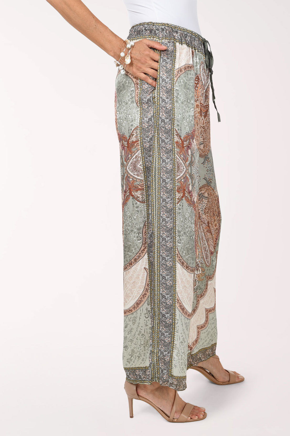 A woman modeling wide-leg patterned pants with a paisley design in muted green, brown, and cream, set against a plain white studio background.